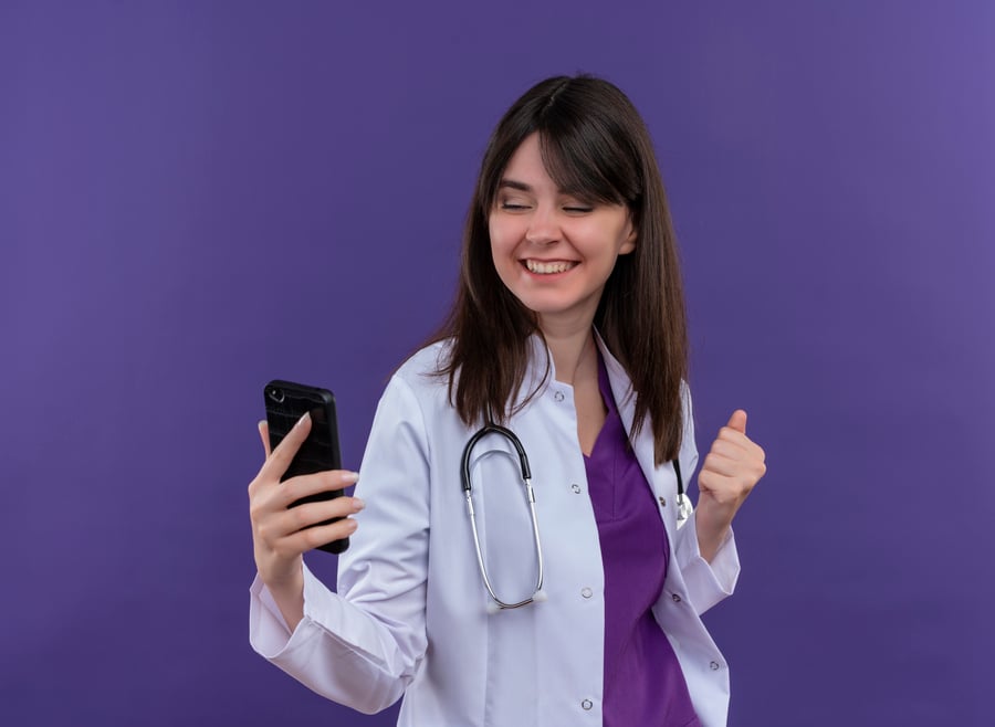 sonriente-joven-doctora-en-bata-medica-con-estetoscopio-sostiene-el-telefono-y-mantiene-el-puno-en-alto-sobre-fondo-violeta-aislado-con-espacio-de-copia (1)